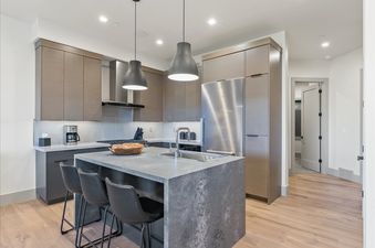 A modern kitchen with a gray stone island, bar stools, stainless steel appliances, and wooden cabinetry.