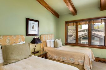 The bedroom in Plaza Lodge 5 Bedroom Gold Unit 7 features two beds with decorative headboards, a bedside table with a lamp, and a large window offering an exterior view of other buildings and mountains.