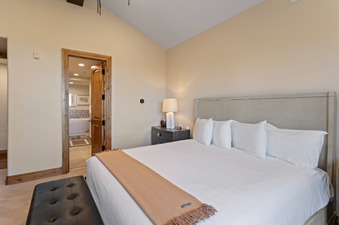 The king-sized bed in the Grand Lodge Five Bedroom Penthouse 406 has a soft, white comforter with a brown throw. The bedroom is decorated in warm, neutral tones and there are wooden doors in the background.