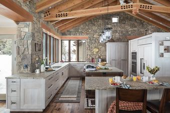A spacious, rustic-modern kitchen at Paintbrush Retreat featuring vaulted wooden beam ceilings, stone walls, light wood cabinetry, granite countertops, modern appliances, and a breakfast bar overlooking a snowy landscape.