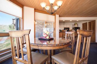 A round wooden dining table with a glass top and four matching chairs, set against a backdrop of large windows with an outdoor view and a glimpse of the kitchen.