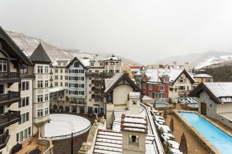 A high-angle view of a charming village nestled in snowy mountains, featuring multi-colored buildings with snow-dusted roofs, a central ice rink, and two outdoor swimming pools.