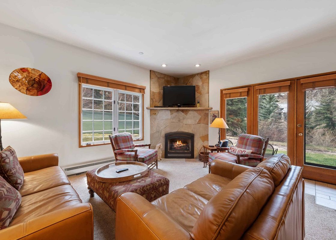 A cozy living room in The Charter E0105 featuring a fireplace, comfortable leather sofas, and plaid armchairs, with large windows overlooking outdoor scenery.