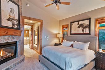 The cozy master bedroom at Rustlers Lodge features a king bed with plush linens, a stone fireplace, and a large window overlooking the surrounding mountains.