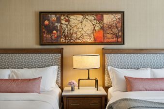 Two queen beds with white linens and red throw pillows are separated by a nightstand with a lamp and vase of flowers, with an autumnal-themed painting above the beds.