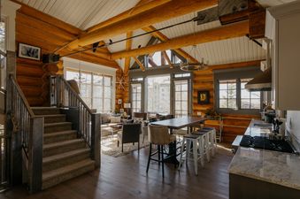 An expansive open-concept living room and kitchen in a luxurious log cabin, featuring large windows, exposed wooden beams, comfortable furniture, a long dining table, and a modern kitchen area.