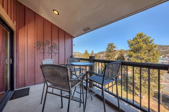 The balcony features a cozy dining set with a table and four chairs, surrounded by warm wood-paneled walls and offering scenic views of evergreen trees and distant mountains.