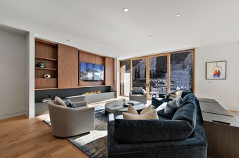 A modern living room at Founders Place 3103 features a fireplace, a TV, blue and gray sofas, and large windows looking out onto a snowy mountain scene with ski lifts.