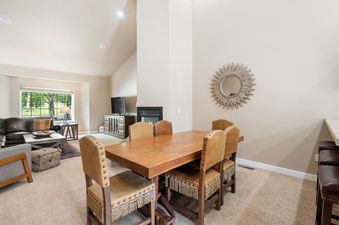 A dining table with six upholstered chairs featuring fringe and nailhead trim is set in an open-concept living space with a fireplace and views of green trees through a large window.