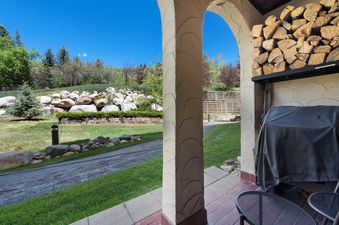 A covered patio with rustic arches, stacked firewood, and a BBQ grill overlooks a green lawn and rock garden at The Charter B1305.