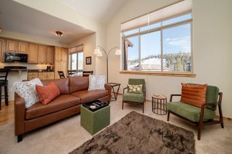 The living area of Trailhead Lodge 731 features a comfortable brown leather sofa, two green armchairs, and a large window overlooking a snowy landscape.