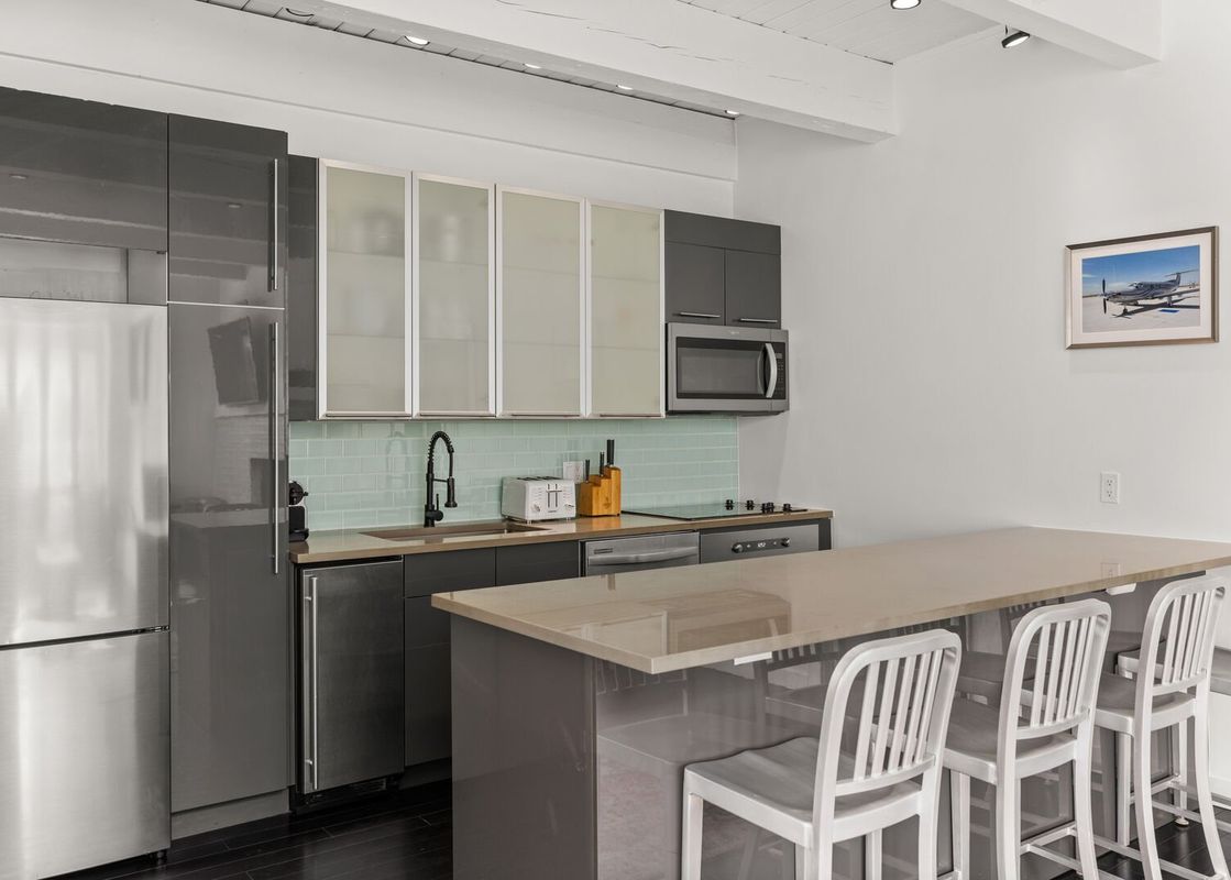 A modern kitchen with stainless steel appliances, grey cabinetry, a light blue subway tile backsplash, and a long kitchen island with four white bar stools.