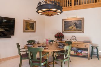 A rustic dining area with a round wooden table, green chairs, and unique chandelier, adorned with art and set for a meal.