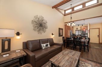 A spacious and inviting living area in Beaver Village 333, featuring a comfortable brown leather sofa, rustic wooden furniture, and a unique metal wall art piece, with a dining area and kitchen visible in the background.