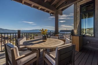 A round wooden dining table with chairs and a BBQ grill on an outdoor patio, overlooking a vast mountain landscape under a clear blue sky.