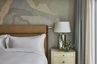 A hotel room with a white bed, brown headboard, a nightstand with a lamp and a vase of flowers, and a window with sheer curtains.