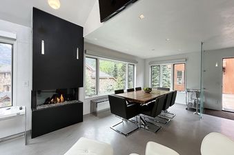 A modern dining room with a fireplace, a large wooden table, and black leather chairs.