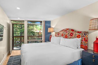 A bedroom with a white bed and a colorful floral headboard, a bedside table with a red lamp, and a window with a view of trees.