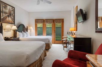 A hotel bedroom with two queen beds, a dresser with a TV, a round table with two stools, and French doors leading to a patio.