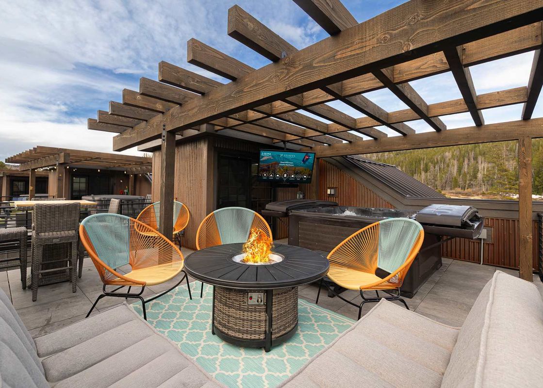 A modern outdoor deck with a fire pit, stylish chairs, and a hot tub, set under a wooden pergola with a view of a forest.