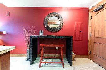 A black vanity table with a red stool in front of it, in front of a red wall with a wooden oval mirror.
