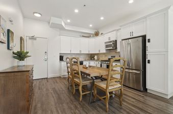 A modern kitchen and dining area at The Mason 1052 featuring white cabinetry, stainless steel appliances, a wooden dining table with four chairs, and a rustic wooden dresser with decorative items.