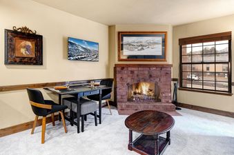 An inviting living and dining space at Copperbottom Inn 202 featuring a fireplace, a flat-screen TV displaying a snowy mountain scene, a dining table, and a window with an exterior view.