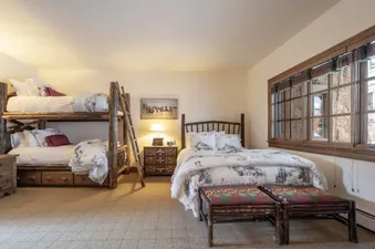 The bedroom in Chalet 12 features a rustic wooden bunk bed and a queen-sized bed, both adorned with white and grey patterned bedding. A nightstand with a lamp sits between the beds, and two upholstered benches are at the foot of the queen bed. A large win