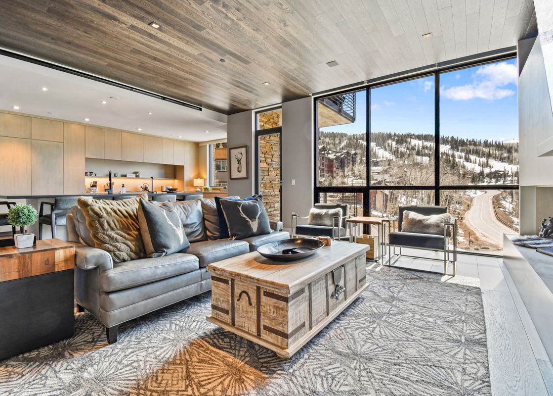 The image shows the inviting great room of One Empire Pass 303, boasting a comfortable seating area, a modern kitchen, and expansive windows that offer stunning views of snow-covered mountains and a scenic road.