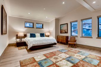 A spacious bedroom in Gondola One Chalet features a queen-sized bed with white linens, blue pillows, bedside tables with lamps, windows offering exterior views, a colorful area rug, a wooden dresser, and a brown leather armchair.