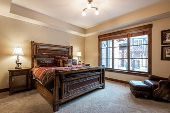 A rustic yet luxurious bedroom with a wooden king bed, two bedside lamps, a large window, and a leather armchair.
