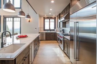A modern kitchen at 7 Lower Cascade Ridge with dark wood cabinetry, stainless steel appliances, and a large island with a sink, all brightly lit by pendant and recessed lighting.