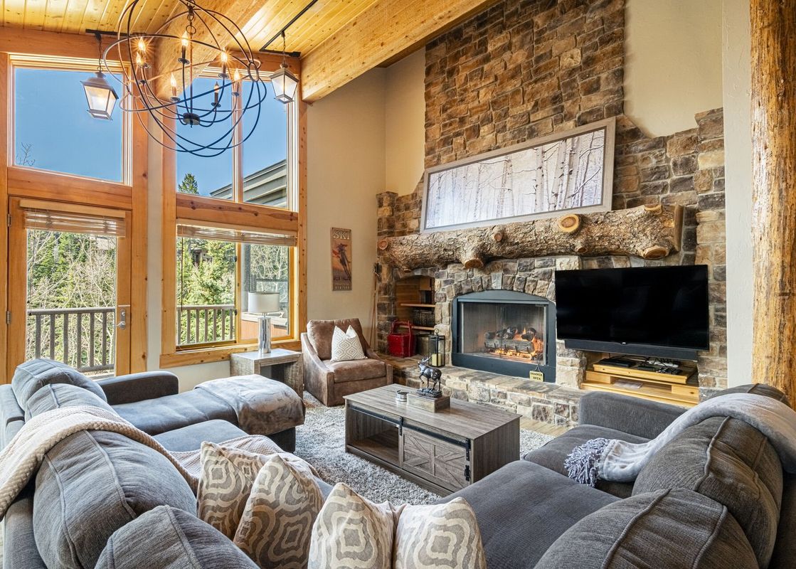 A cozy and elegant living room in Royal Plaza 403 features a stone fireplace, comfortable gray sectionals, a rustic coffee table, and large windows with scenic outdoor views.