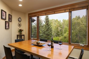 A dining area with a large wooden table and black chairs, featuring a bottle of wine and glasses, with a large window overlooking a lush green forest at Terracehouse 63.
