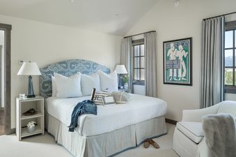 A serene bedroom at Four Pines 12 features a blue patterned headboard, white and blue bedding, and a winter sports painting, with mountain views from the windows.