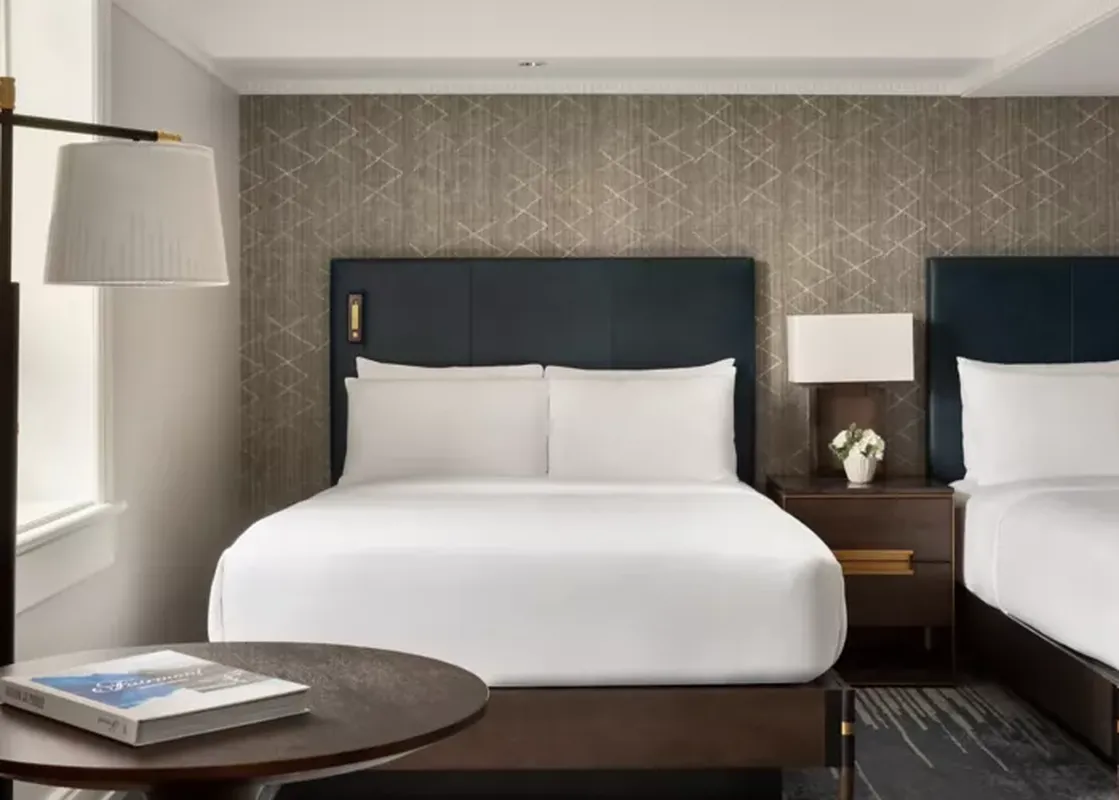 A sophisticated bedroom at Fairmont Banff Springs Gatehouse 2 Queens, featuring two queen beds with navy headboards, white linens, a textured accent wall, and elegant lighting.