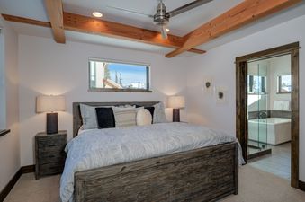 A cozy bedroom in Homestead Chalet 8 Claim Jumper features a rustic king-size bed with a patterned duvet, two nightstands with lamps, a window with mountain views, and an ensuite bathroom with a white bathtub.