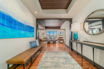 The entryway of Lumiere Two Bedroom Residence, featuring a long leather bench and a large painting on the wall. The hallway opens up into a bright living area with a large window showcasing mountain views.