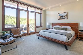 A primary bedroom with a large wooden bed, two leather armchairs, and a private balcony overlooking lush green mountains at Victoria Condominiums 2 Bedroom.