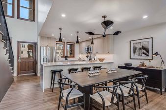 A modern open-plan kitchen and dining area in Woodrun Place 56 featuring light wood cabinets, a kitchen island with bar stools, a dark wood dining table with black chairs, and a contemporary ceiling fan.