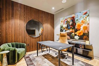 A modern office space in Founders Place 2502 with a wooden accent wall, a circular mirror, a dark desk, a tan office chair, a green armchair, and a large, colorful floral painting.