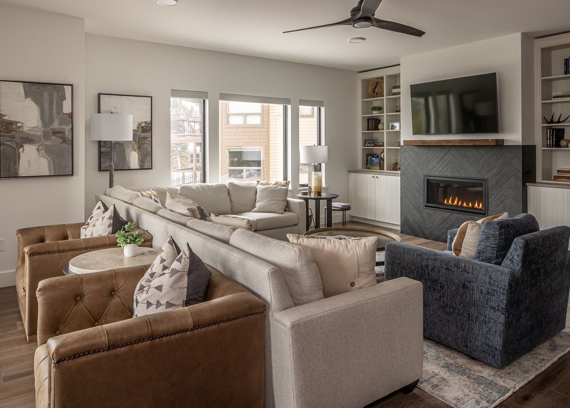 The living area of Beaverhead Condo 301 features a cozy fireplace, ample seating with light-colored sectionals and dark leather armchairs, and large windows offering exterior views.
