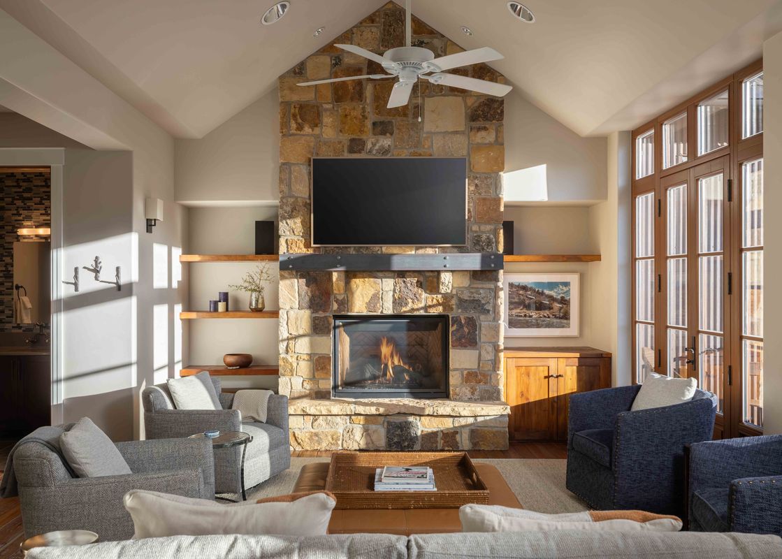 A living room with a stone fireplace, a ceiling fan, and a large window with an exterior view.