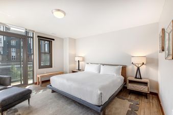 A modern primary bedroom at Lowell 402B featuring a king-sized bed, two nightstands with lamps, a balcony, a wooden bench, and a dark upholstered chaise lounge.