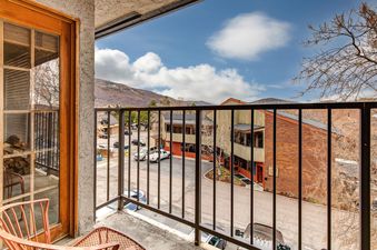 A balcony with a metal railing offers a view of mountain landscapes, buildings, and a clear blue sky with clouds.