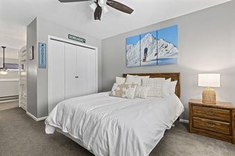 A white, king-sized bed sits in the center of a bedroom in Wolf Lodge Condo 1412,  with a wooden headboard and a white duvet.  The room has a gray wall and wooden nightstand.  The room also has a pair of white closet doors with a sign that reads "Snowboar