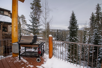 A stainless steel BBQ grill sits on a snowy wooden deck, offering views of a snow-covered forest and distant mountains under a cloudy sky.