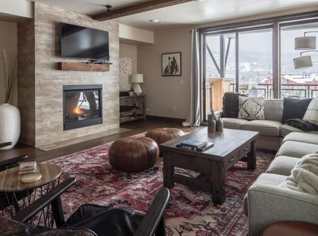 The cozy living area of Mountain Lake Condo 401B features a stone fireplace with a mounted TV, plush sectional sofas, a wooden coffee table, and large glass doors opening to a balcony with mountain views.