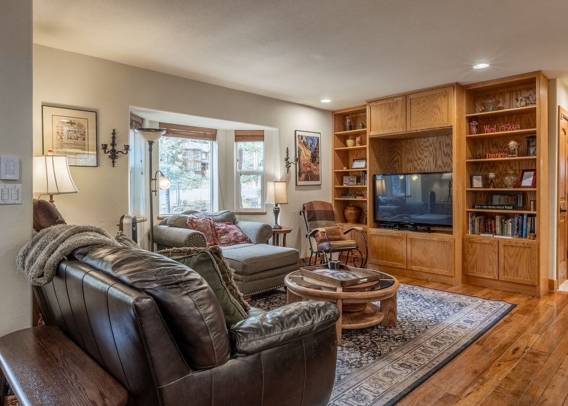 The inviting living room features a dark leather sofa, a chaise lounge, a wooden coffee table, and a built-in entertainment center with a television and shelves, all illuminated by warm lighting and natural light from the bay windows.