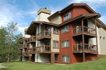A multi-story building with reddish-brown and light beige siding, featuring numerous balconies and surrounded by a green lawn, walking path, and trees under a clear blue sky, showcasing the exterior of the Ranch at Steamboat RA303.
